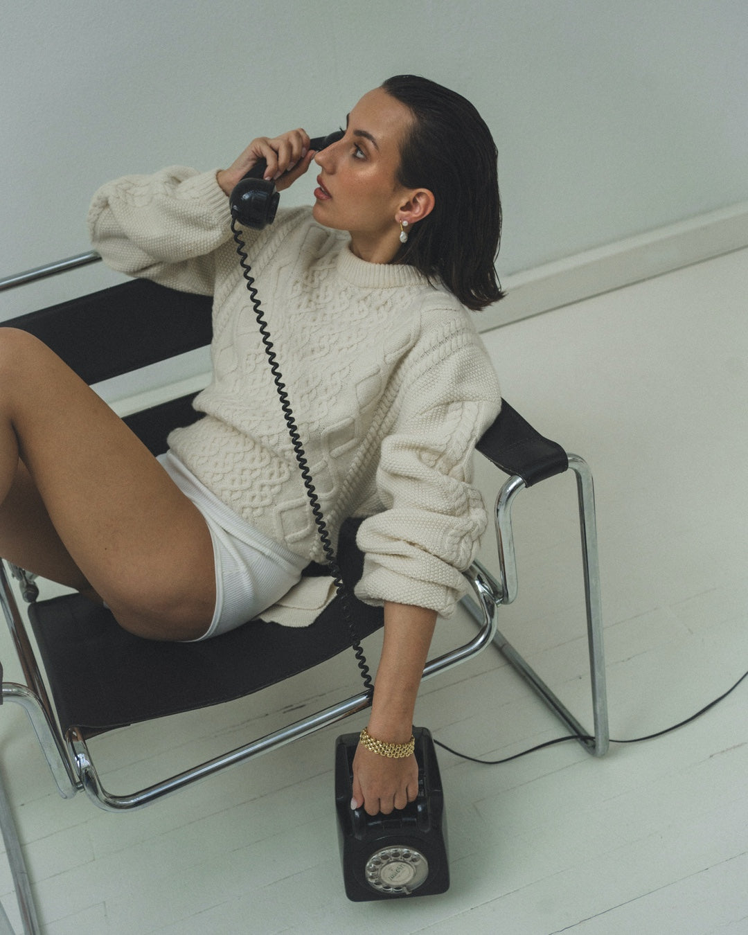 Woman with black phone sitting on a chair wearing a sweater and shorts, showcasing natural versus cultured pearls jewelry.