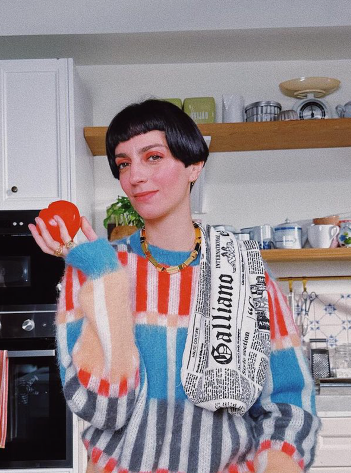 A person holding a red object while wearing a stylish colorful sweater and a bag, posing in a kitchen.