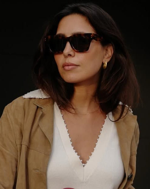 Woman wearing sunglasses and gold earrings with a stylish beige jacket.