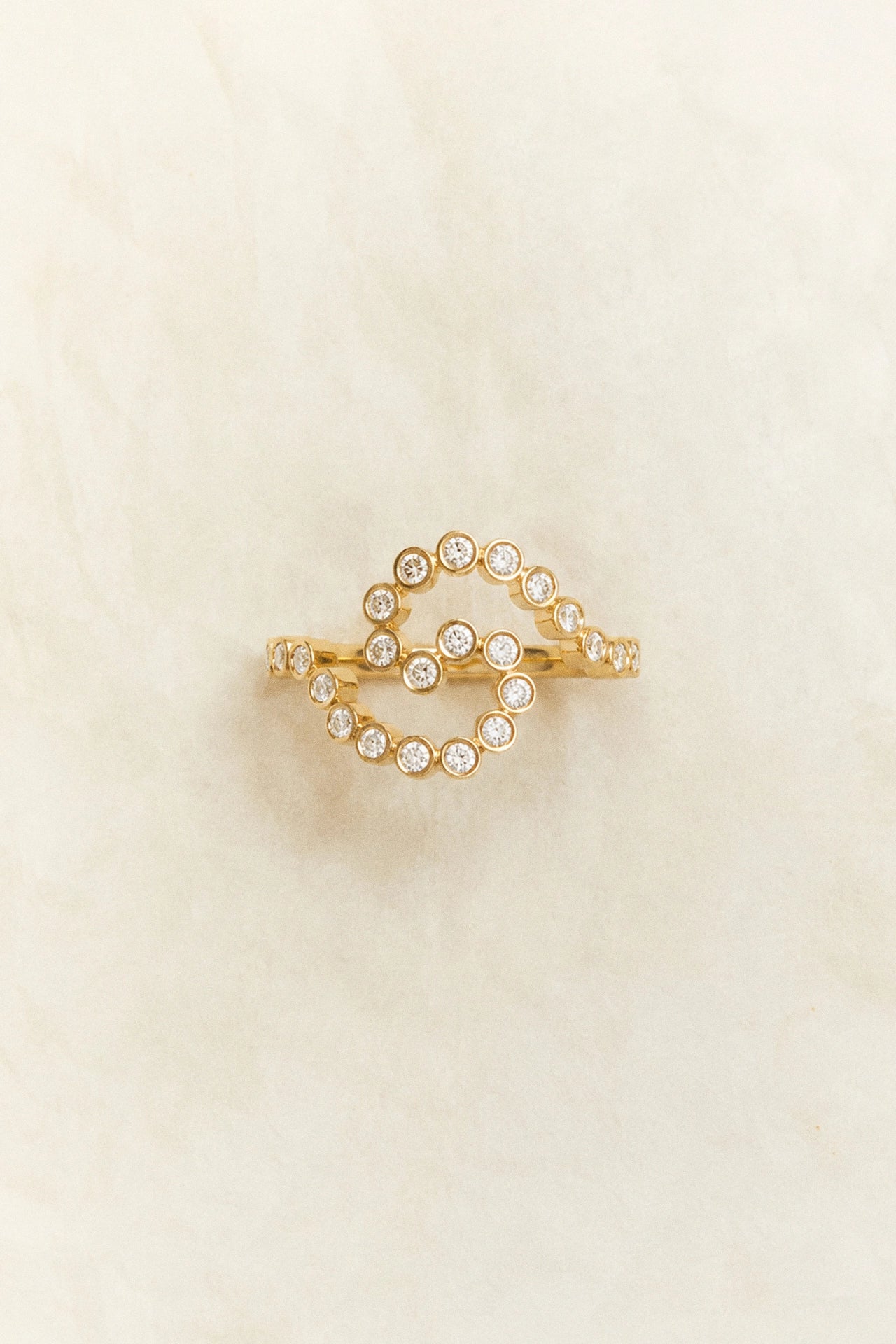 A gold ring featuring a double-loop design set with small clear round gemstones, displayed on a soft, textured cream background.