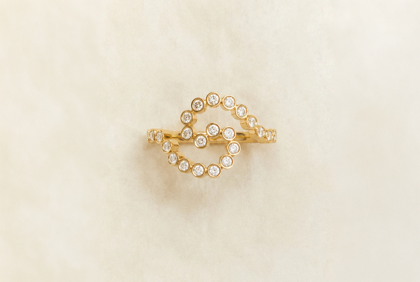 A gold ring featuring a double-loop design set with small clear round gemstones, displayed on a soft, textured cream background.
