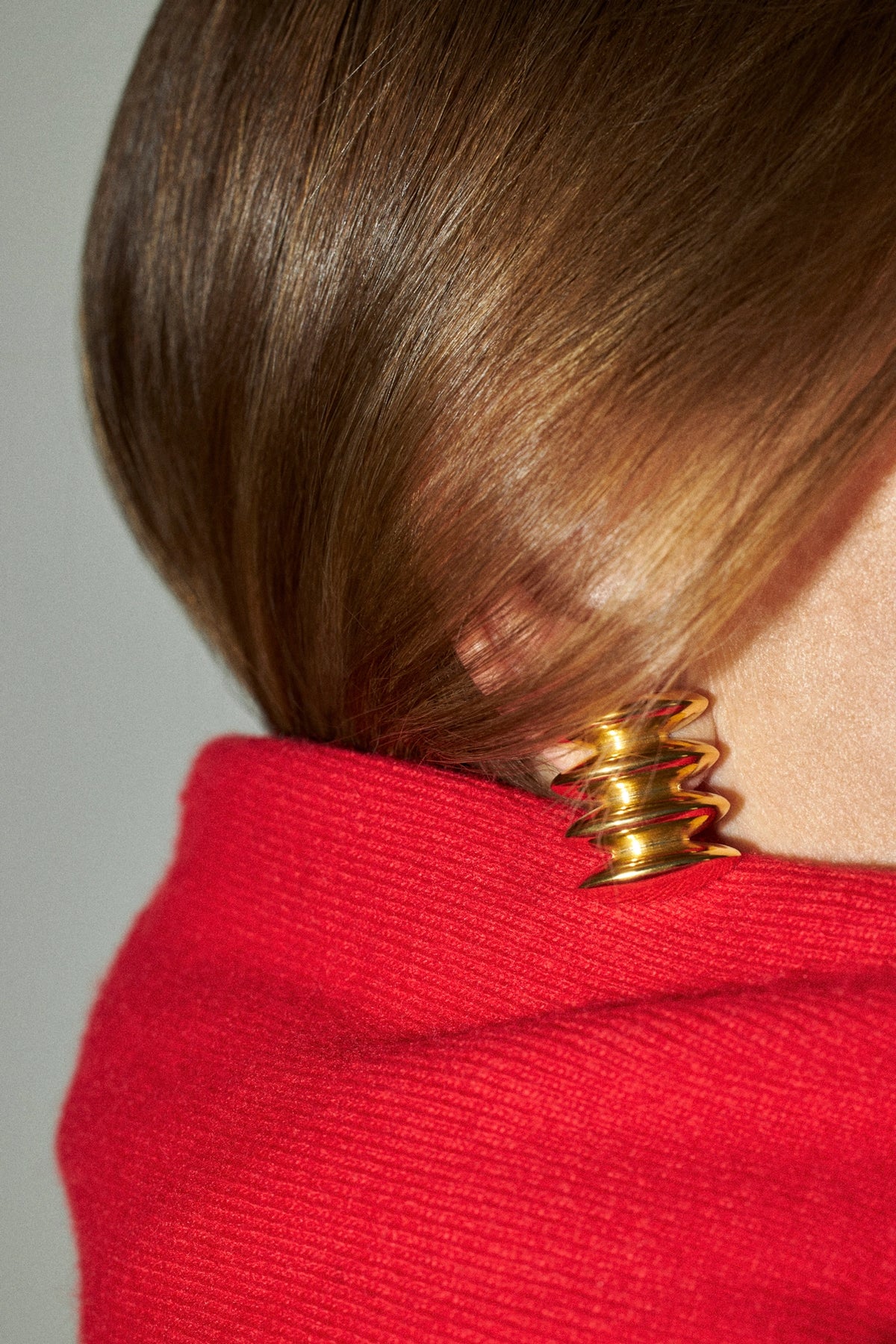 A close-up shot of a person's brown hair tied back with a distinctive gold coiled spring hair accessory, wearing a bright red knit top against a neutral gray background.