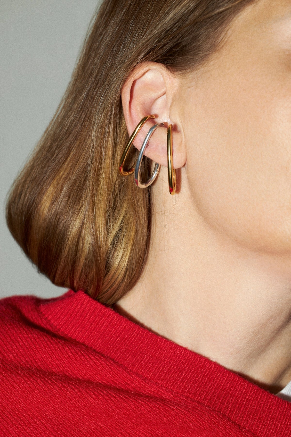A close-up of a model's ear wearing three stacked, thin hoop earrings in different metallic finishes, including gold, rose gold, and silver, paired with a red knit top against a neutral gray background.