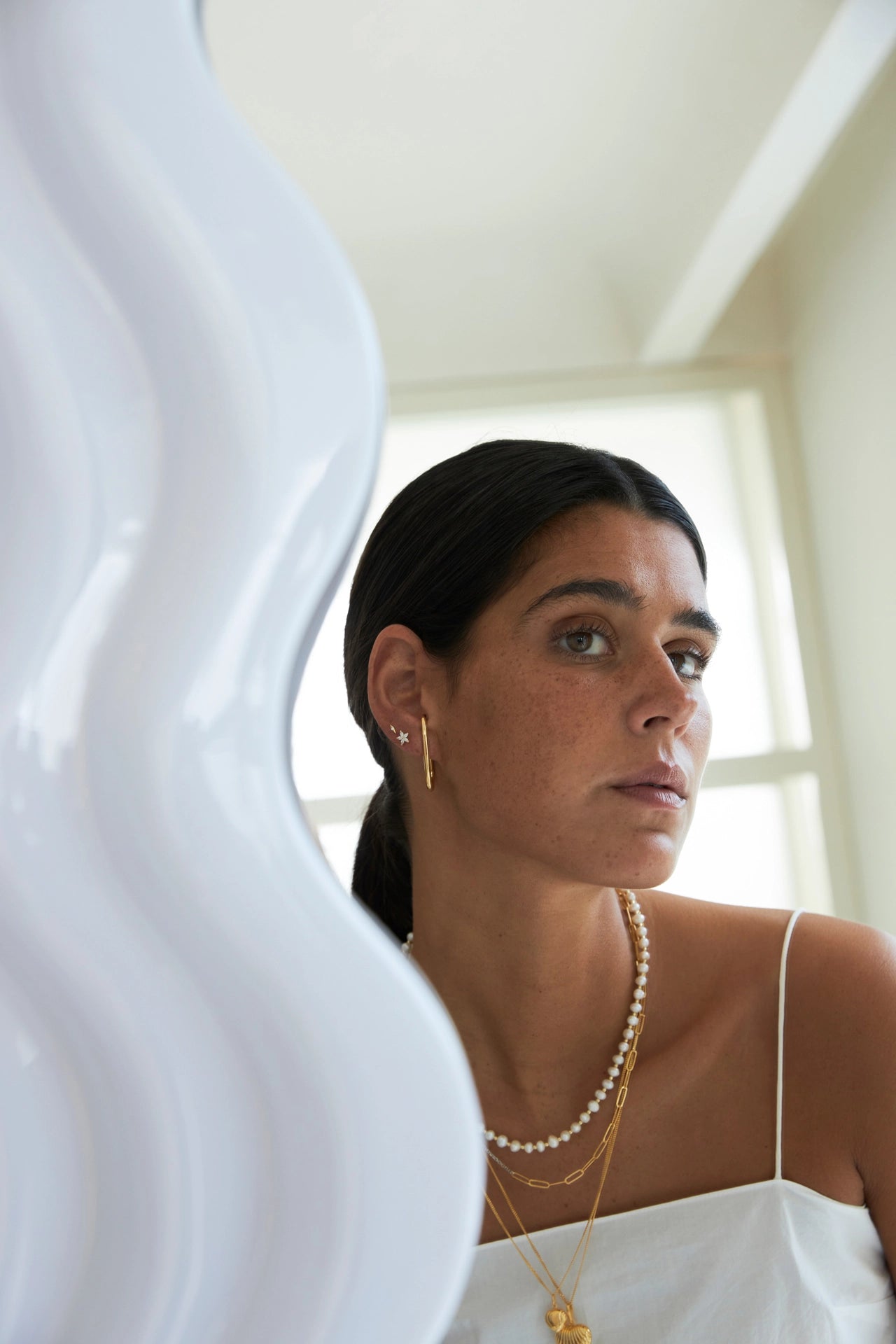 Woman wearing gold earrings and necklaces in a softly lit room.