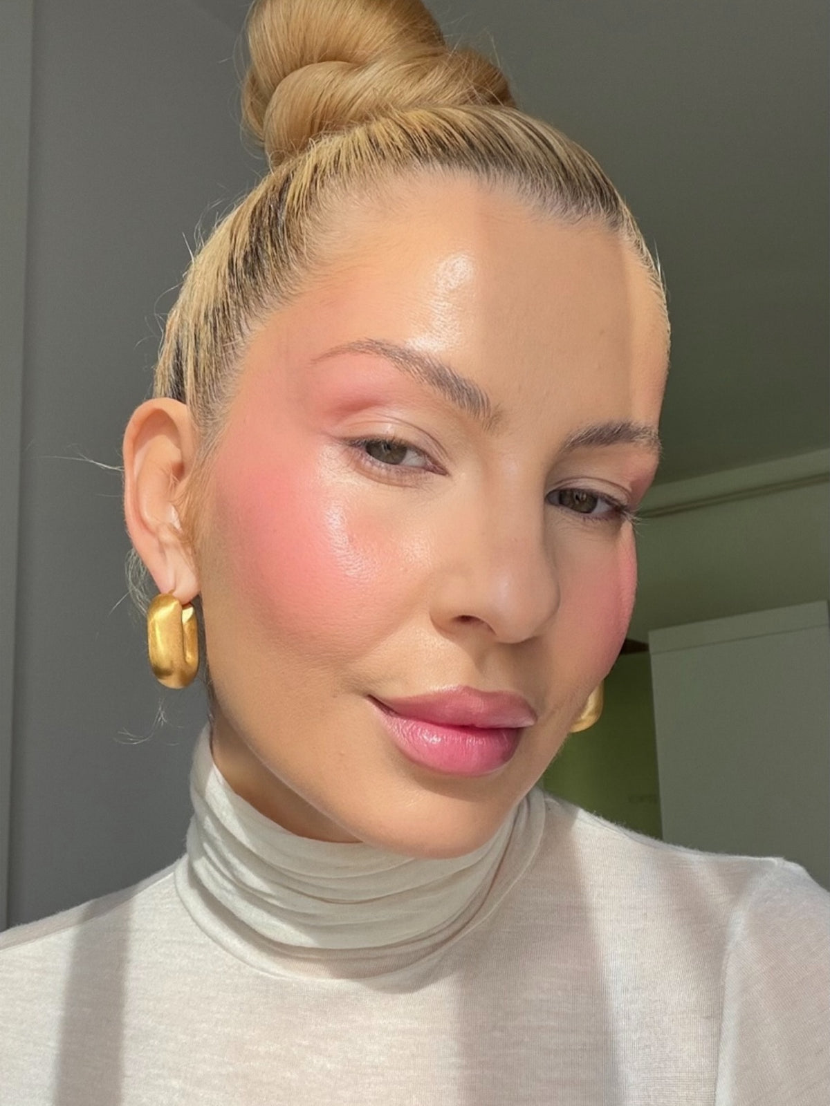 Close-up of a woman wearing golden earrings, with a soft pink blush and glossy lips in natural light.