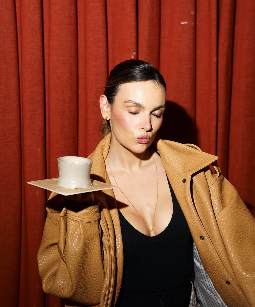 A woman with a stylish leather jacket poses with a ceramic cup on a plate against a red curtain.