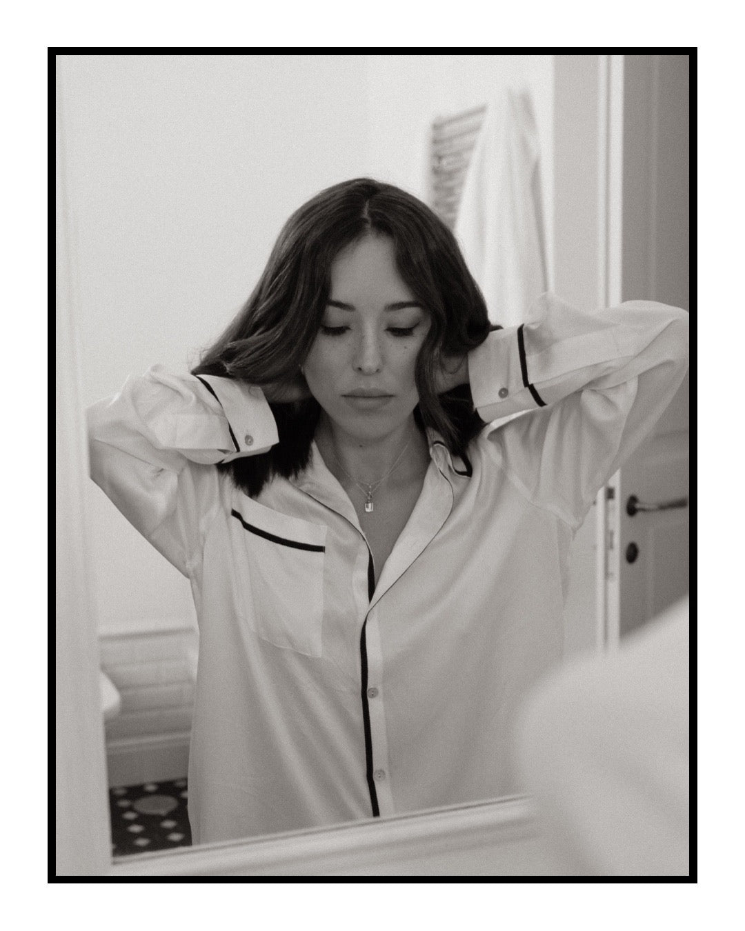 Woman adjusting her hair in front of a mirror wearing a white shirt with black trim.