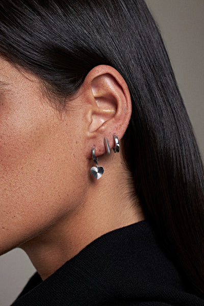 A close-up side profile of a model with dark hair wearing a pair of silver hoop earrings with dangling heart charms.